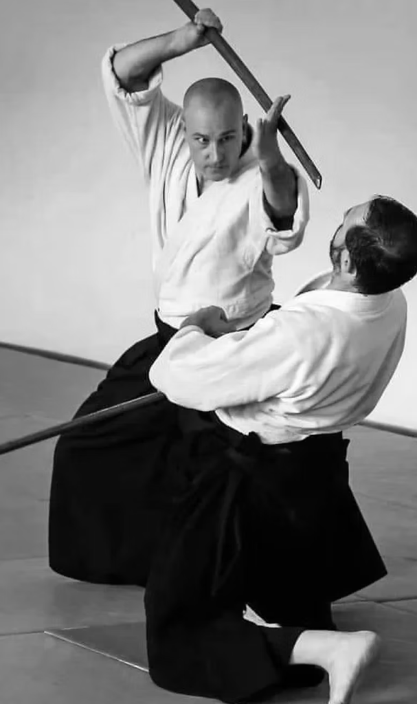 A black and white photo of two martial artists practicing with wooden swords. One is leaning backwards, posture broken, with the other driving down the point of the sword at first's head.