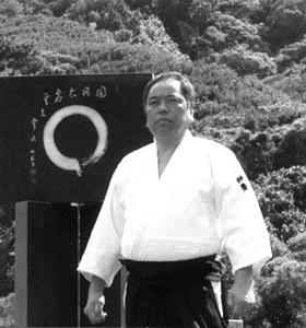 A black and white photo of a stout Japanese man in traditional Aikido attire, standing in front of a wooden screen painted with a Zen enso (circle drawn with a single brush-stroke).