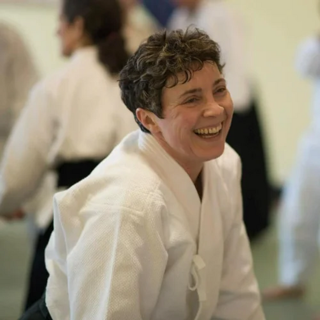 A picture of a woman in a martial arts uniform. The woman is leaning forward and smiling with other martial artists in a blurred background.